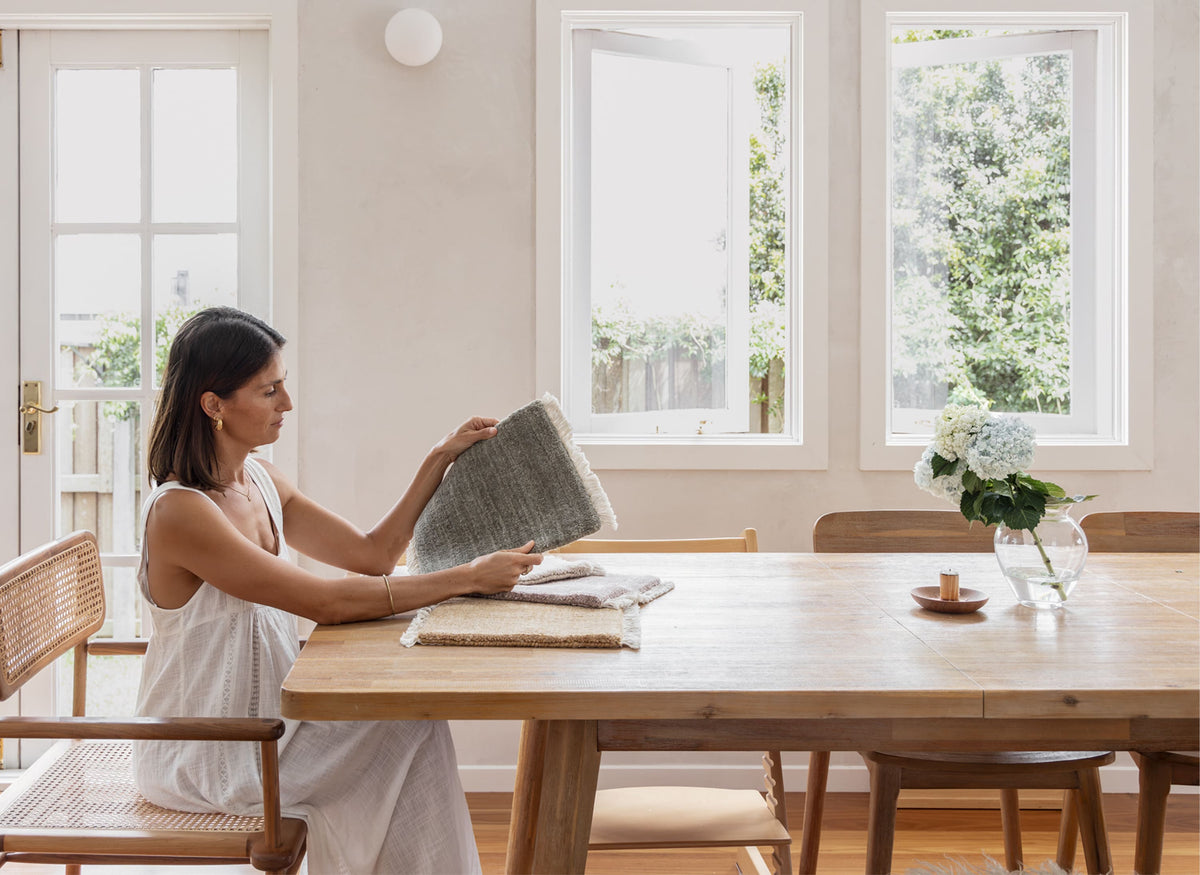 Woman holding a Bhakti rug sample in a bright, naturally lit room with wooden furniture.