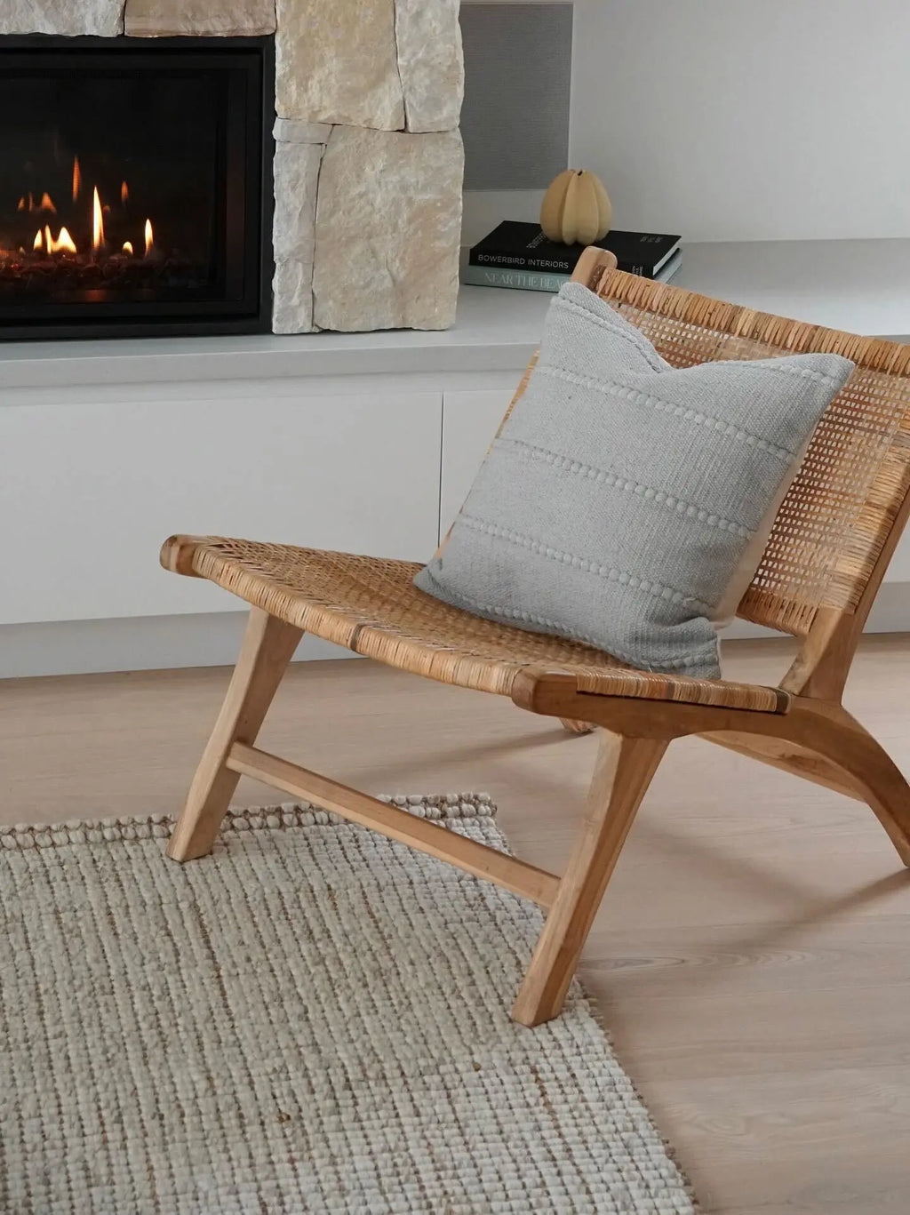 A neutral wool and jute natural fibre rug underneath a wicker chair and grey pillow in front of a stone fireplace.