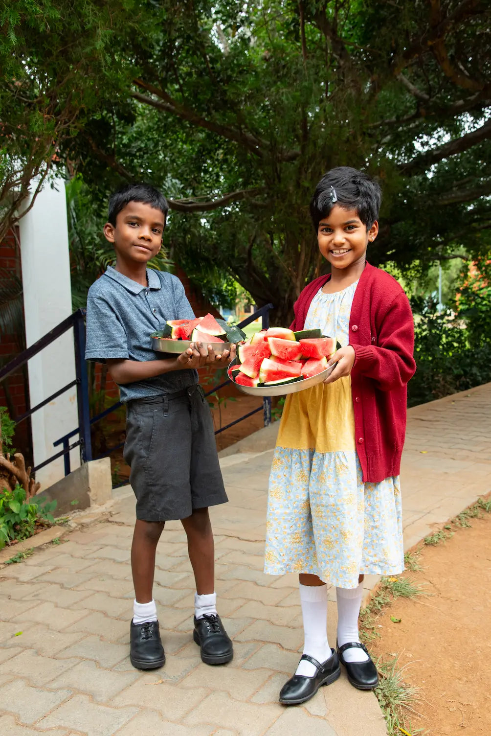 Two children holding plates of watermelon outdoors on a path with trees in the background.
