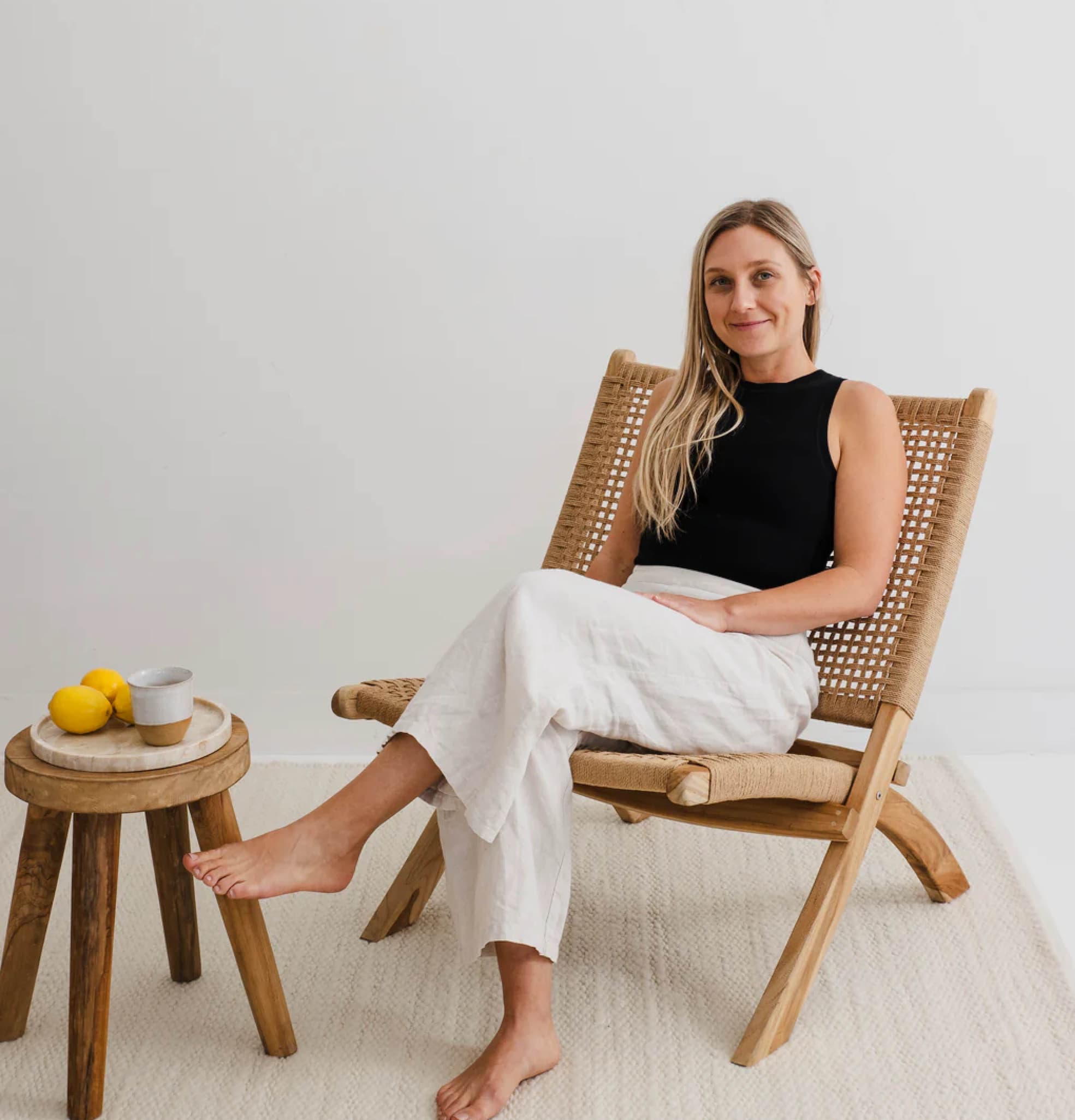 Woman sitting on a wooden chair in a minimalistic room with a small table and lemons. Woman is Jess Founder of Rugs for Good.