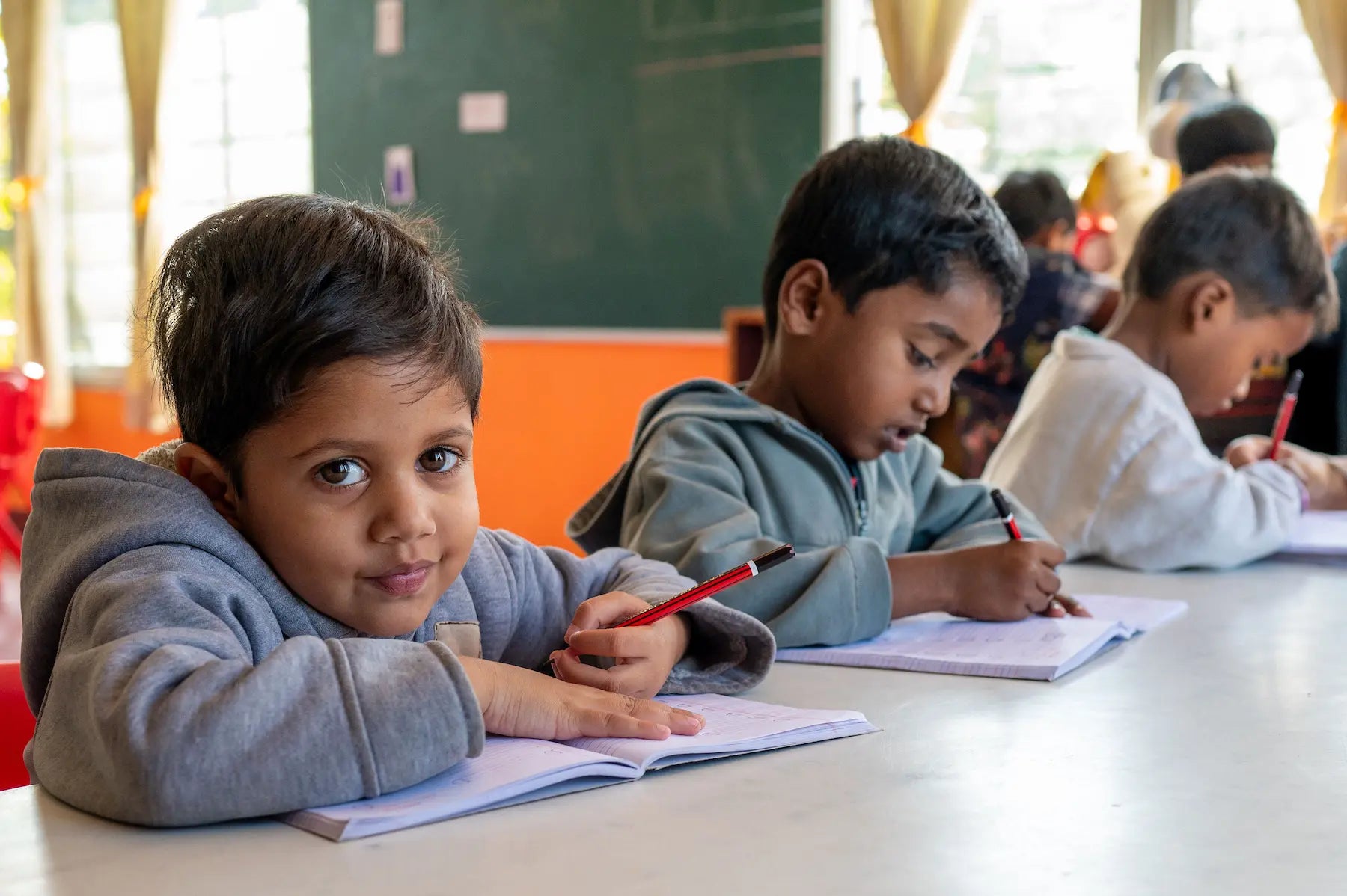 Children sitting at a table in a classroom, writing in notebooks with red pens.